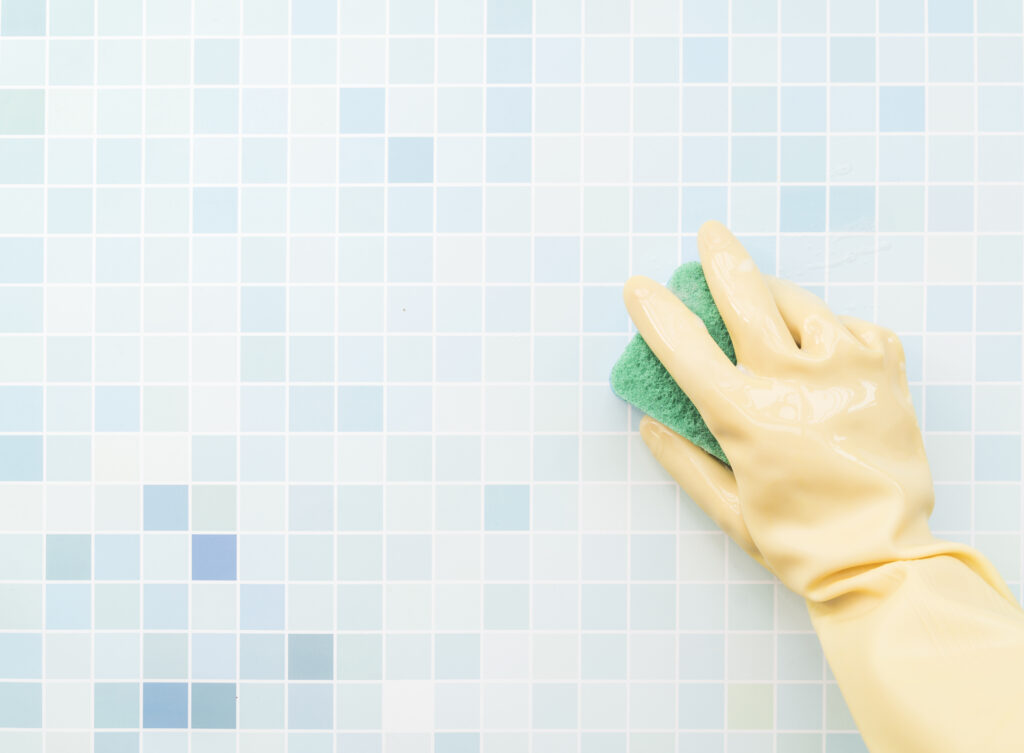 hand in yellow glove scrubbing blue bathroom tiles with a green sponge